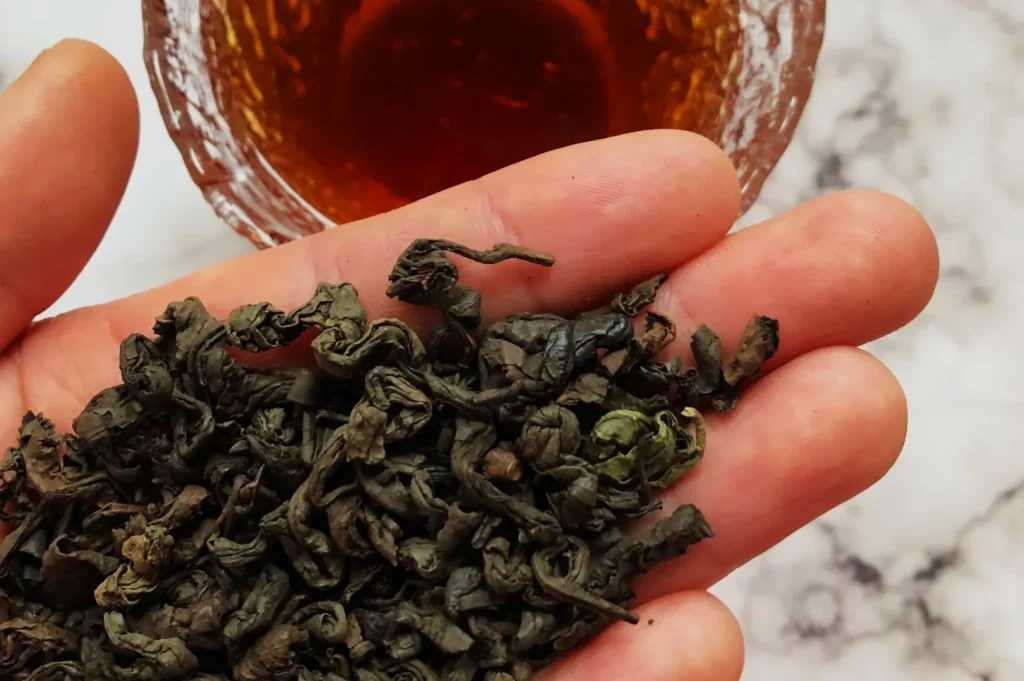 A hand holding dried green tea leaves with a cup of brewed tea in the background.