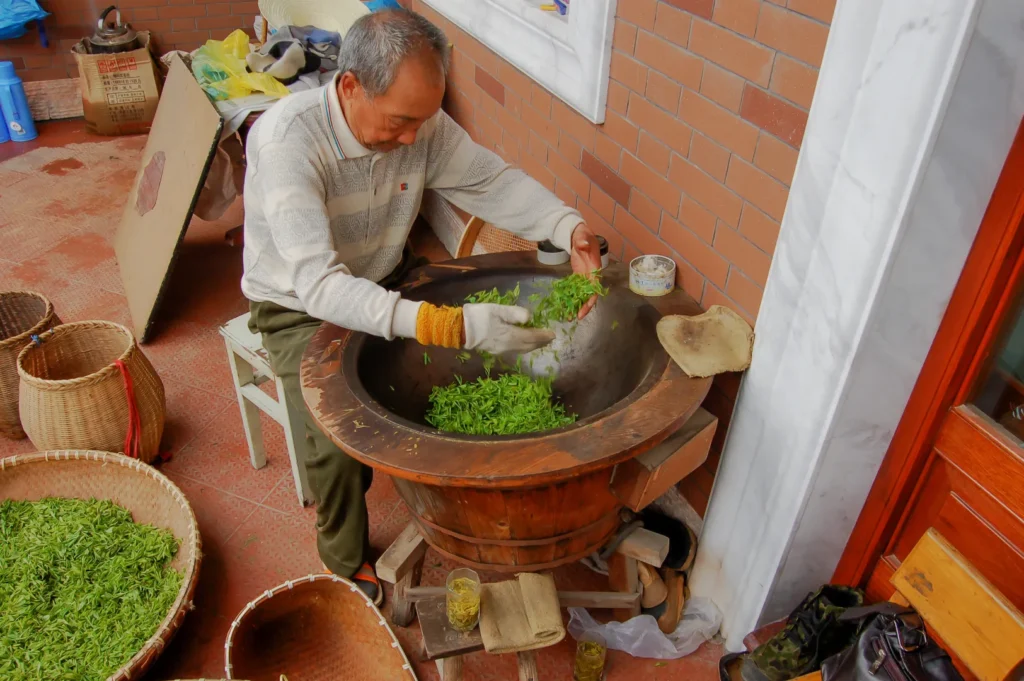 A man in a sweater working with green tea leaves in a large wooden bowl, surrounded by baskets.