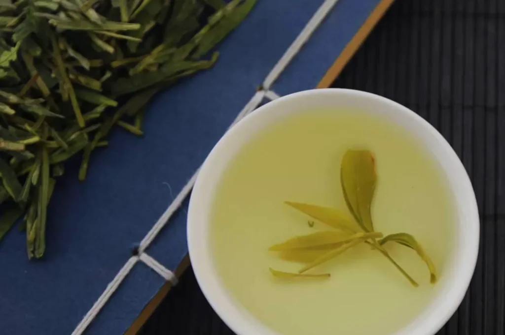 A cup of light green tea with a sprig of tea leaves next to it, placed on a blue background.