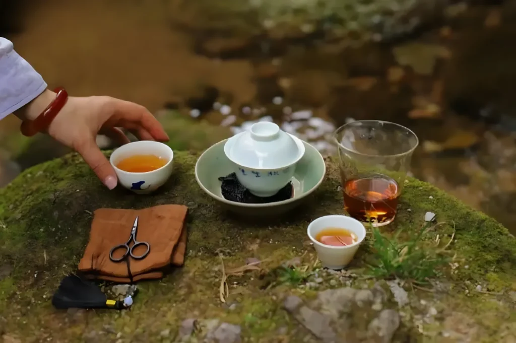 A hand reaching for a small cup of tea beside a teapot, a bowl, and a glass of tea on a stone surface near a stream.