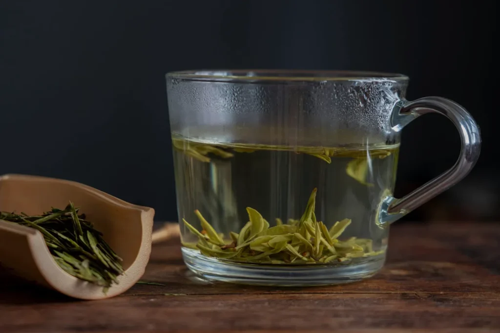 A glass cup filled with green tea and tea leaves, with a small scoop of loose tea leaves beside it.