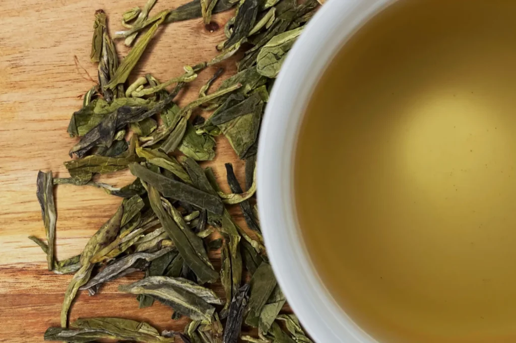 A cup of green tea surrounded by loose green tea leaves on a wooden surface.
