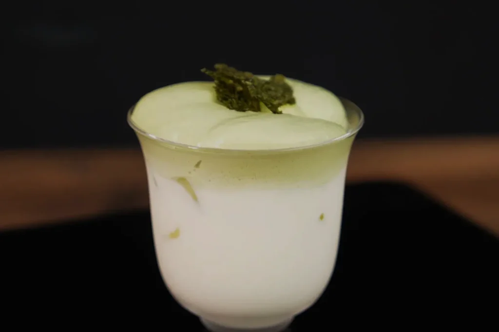 A dessert in a glass, featuring a creamy white base with a green layer on top and topped with some leafy garnish.
