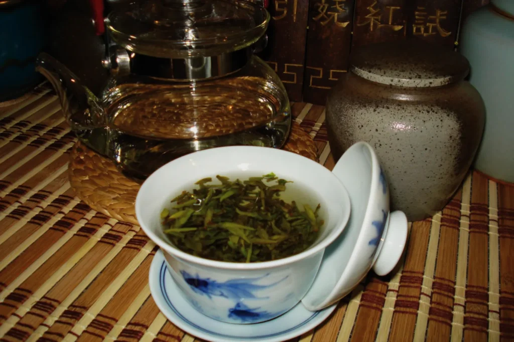 A cup of tea with loose green tea leaves, a teapot in the background, and a decorative pot on a bamboo mat.