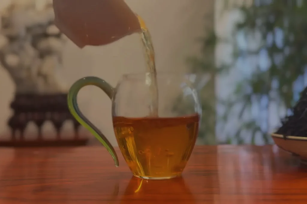 A transparent glass teapot pouring tea into a glass cup on a wooden table.