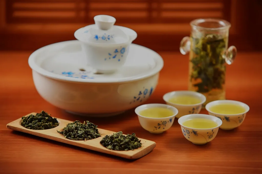 A traditional tea set with a teapot, teacups, and two types of loose tea leaves displayed on a wooden tray, alongside a glass container of brewed tea.