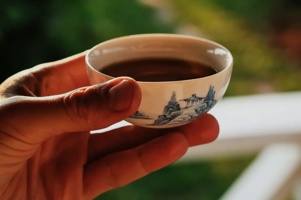 A hand holding a small ceramic cup with a scenic blue and white design, filled with tea.