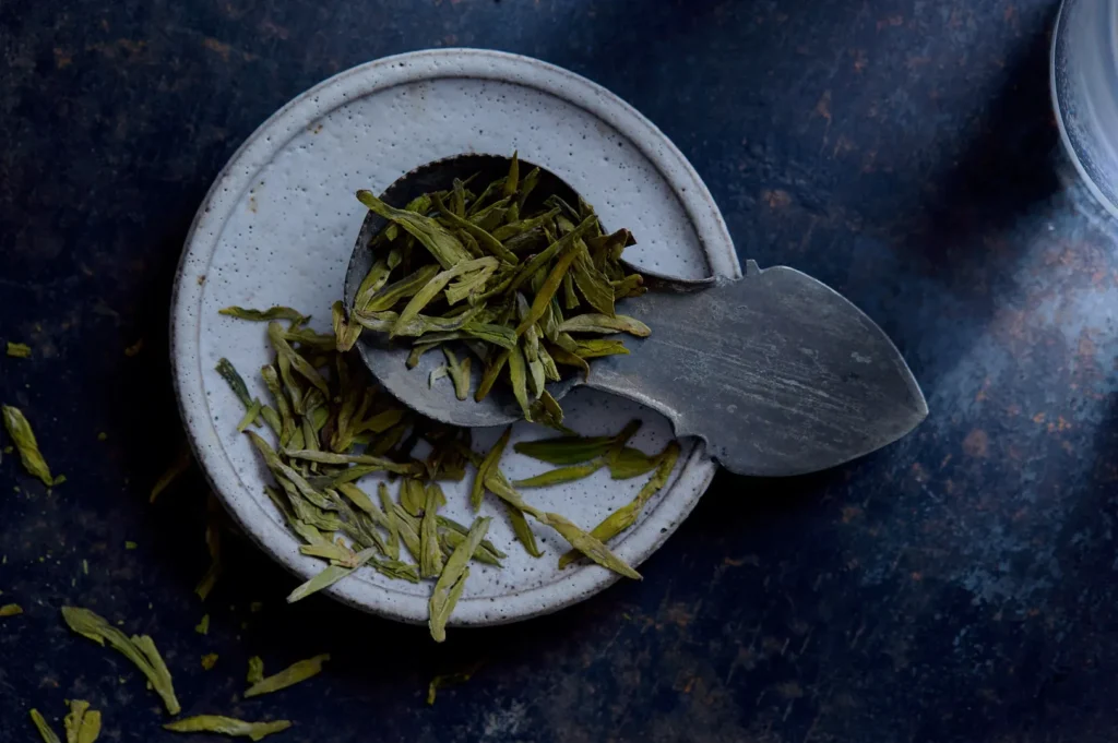A small dish with a metal spoon holding loose tea leaves on a textured surface.