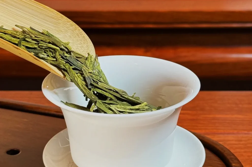 A wooden scoop pouring dried green tea leaves into a white teacup.