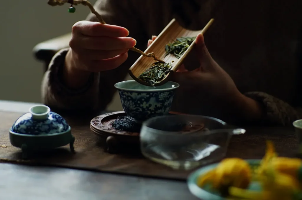 A person preparing tea, using a bamboo scoop to add tea leaves to a ceramic cup, with tea utensils and a small plate of food on a wooden table.