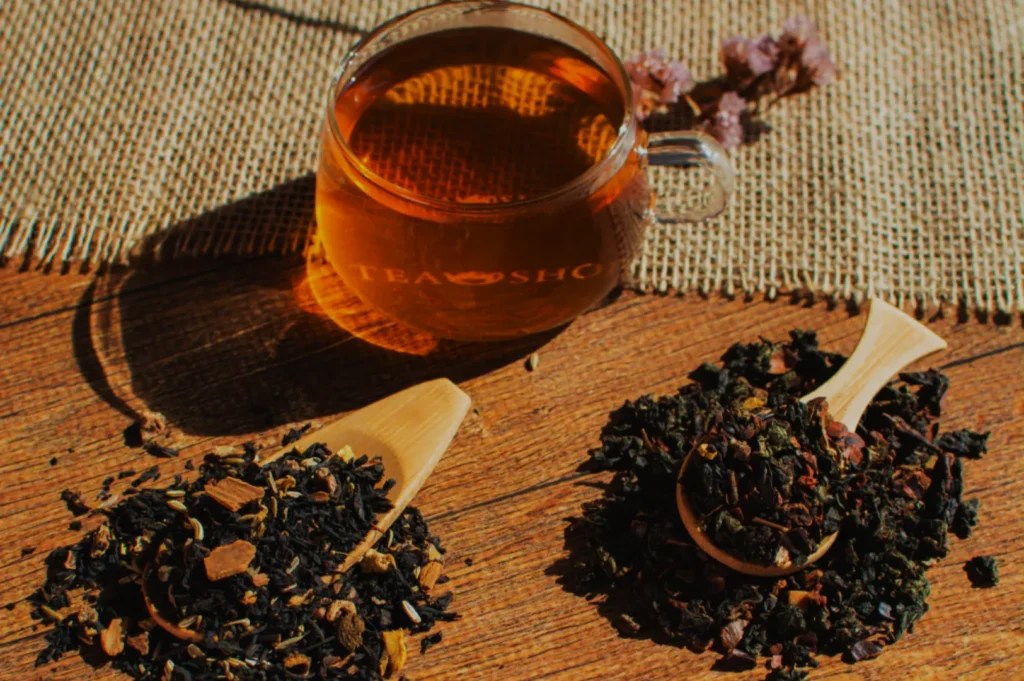 A cup of tea surrounded by two wooden spoons filled with loose tea leaves, on a textured surface with a burlap background.