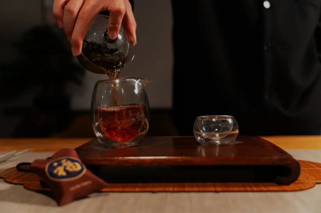 A person pouring tea from a pot into a glass cup on a wooden tray, with a small transparent cup beside it.