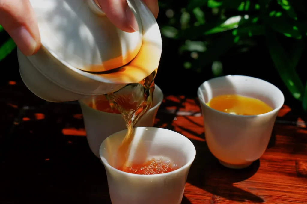 A hand is pouring tea from a teapot into small, white cups, with other cups visible in the background surrounded by greenery.