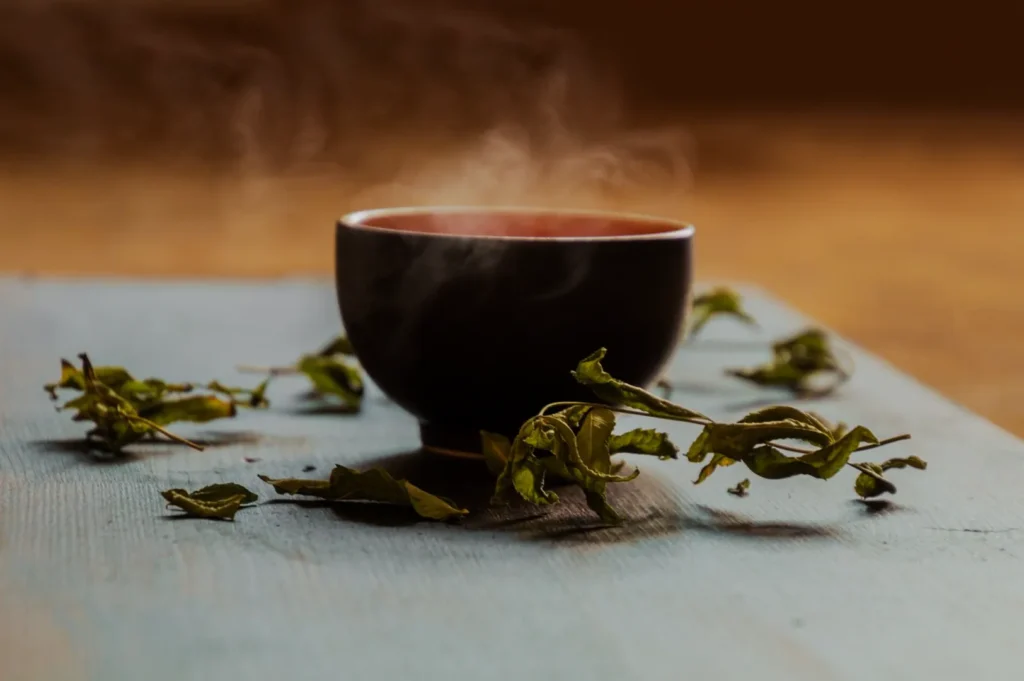 A steaming cup of tea with dried leaves scattered around on a wooden surface.