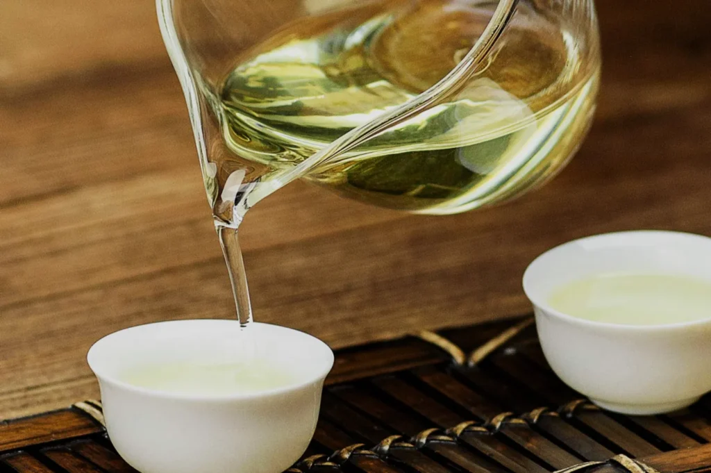 A glass teapot pouring green tea into small white cups on a bamboo mat.