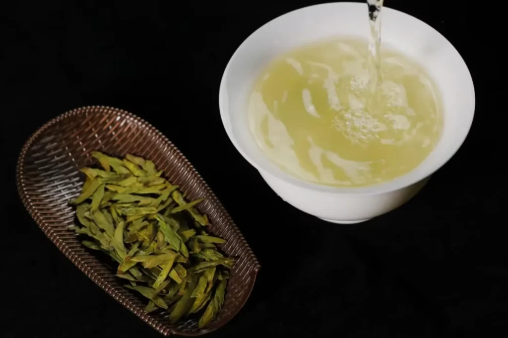 A bowl of light-colored tea being poured next to a woven dish of green tea leaves.