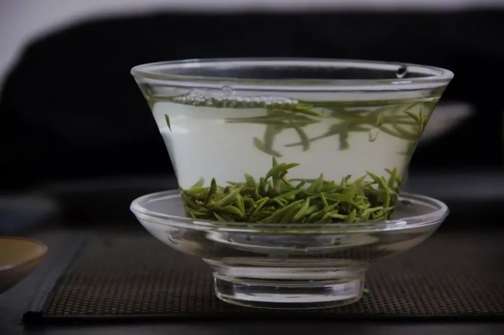 A glass teacup with green tea and tea leaves floating in it, placed on a small glass saucer.