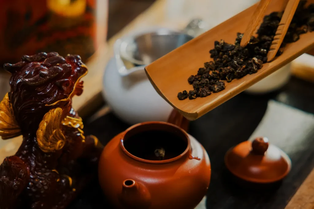 A wooden scoop pouring dried tea leaves into a small teapot, with a decorative dragon figurine and a white teapot in the background.