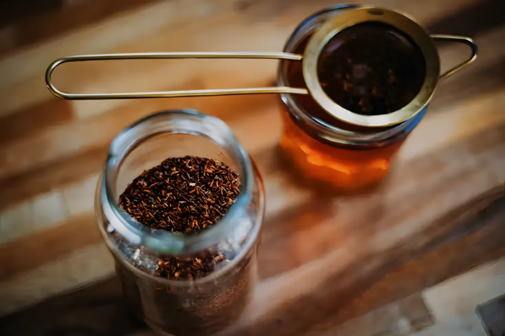 A glass jar filled with loose tea leaves next to another jar with a strainer above it, both sitting on a wooden surface.