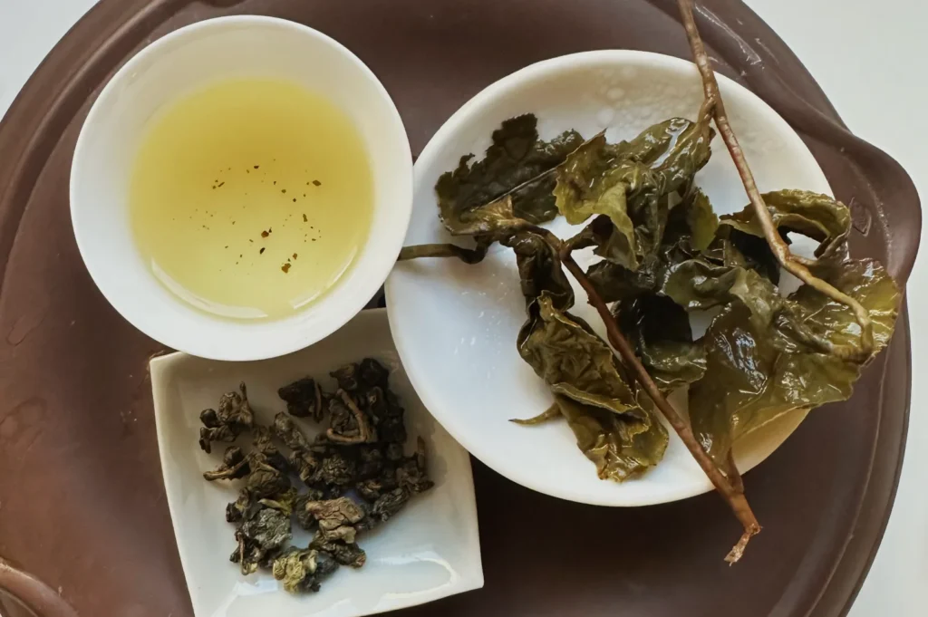 A tray with a cup of light yellow tea, dried tea leaves, and a small dish of dark green tea leaves.