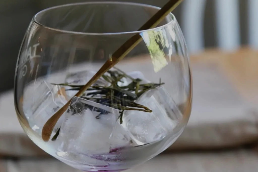 A glass bowl filled with ice and garnished with green herbs, with a stir stick resting on top.