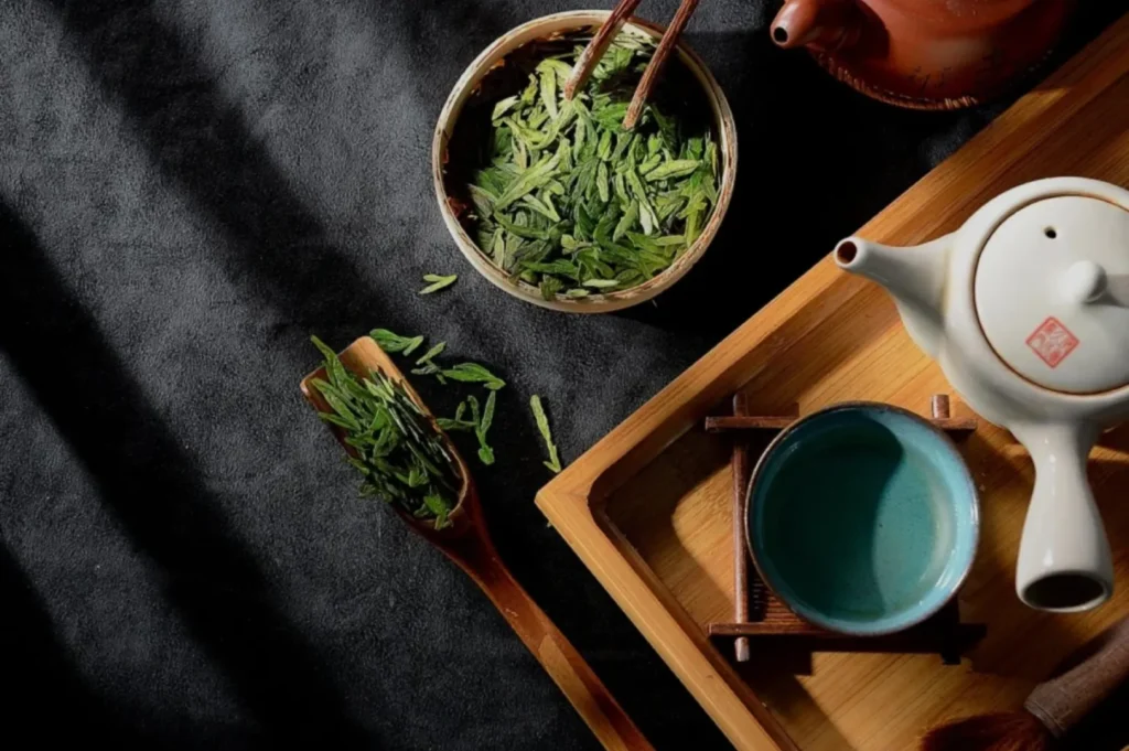A flat lay of green tea leaves in a bowl and on a wooden spoon, with a teapot and a cup on a wooden tray, set against a dark background.