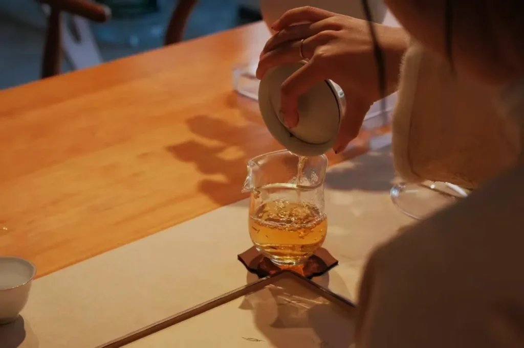 A person pouring tea from a teapot into a glass cup on a wooden table.