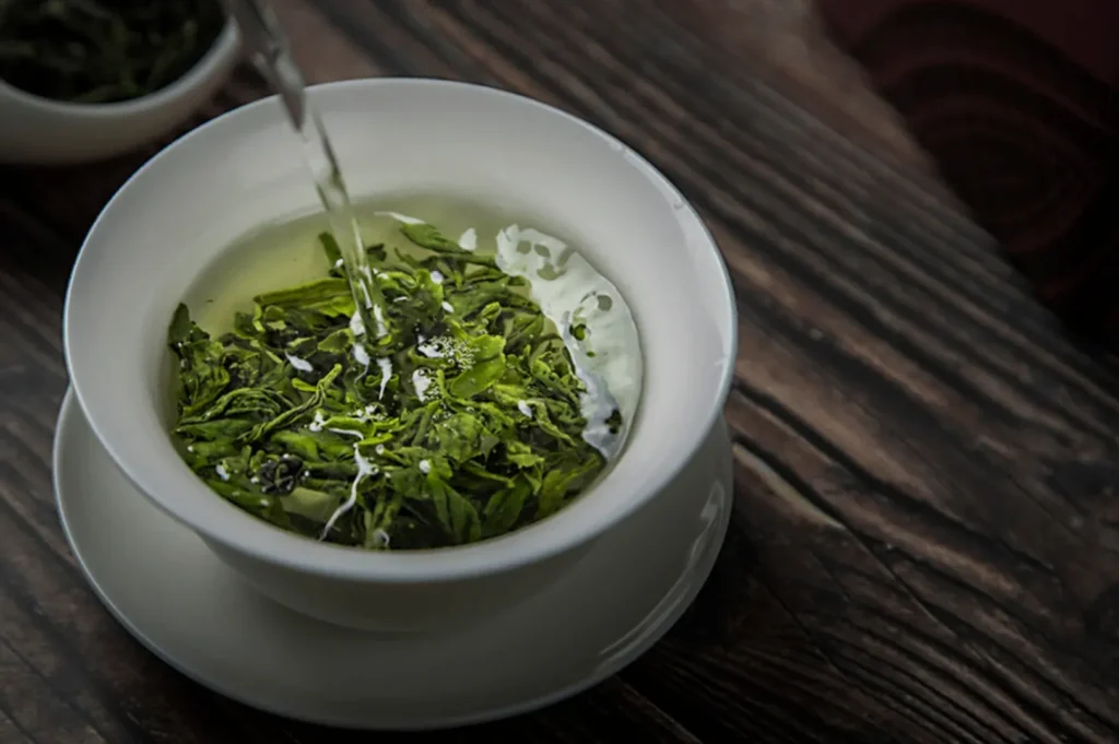 A close-up of green tea being poured into a bowl with vibrant green tea leaves floating on the surface, set on a wooden table.