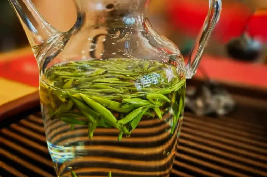 A glass pitcher filled with water and green tea leaves, sitting on a wooden surface with a decorative background.