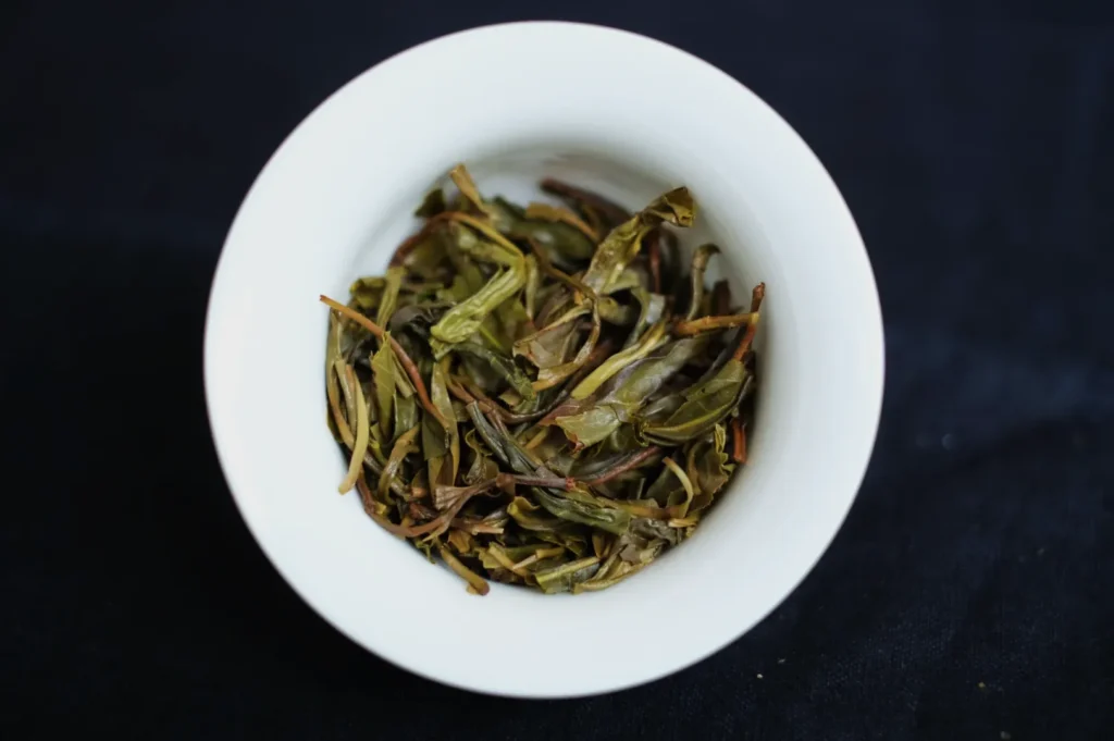A small white bowl filled with dried green tea leaves on a dark background.