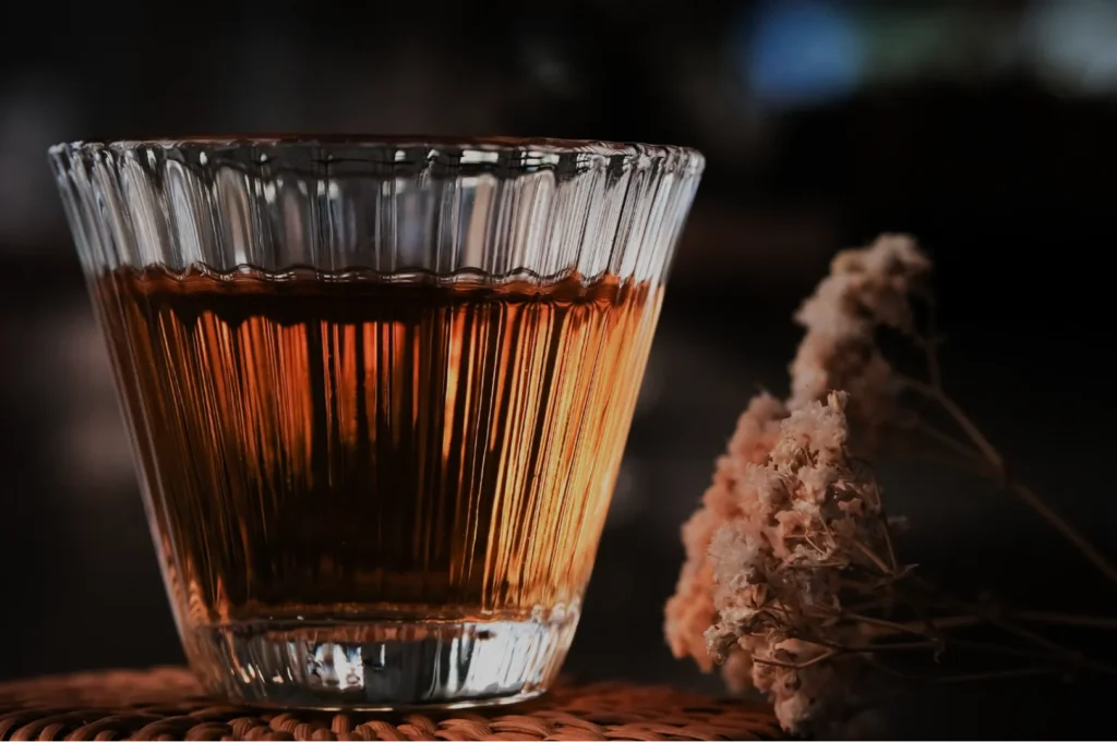 A glass of amber-colored liquid on a textured surface, with dried flowers in the foreground.