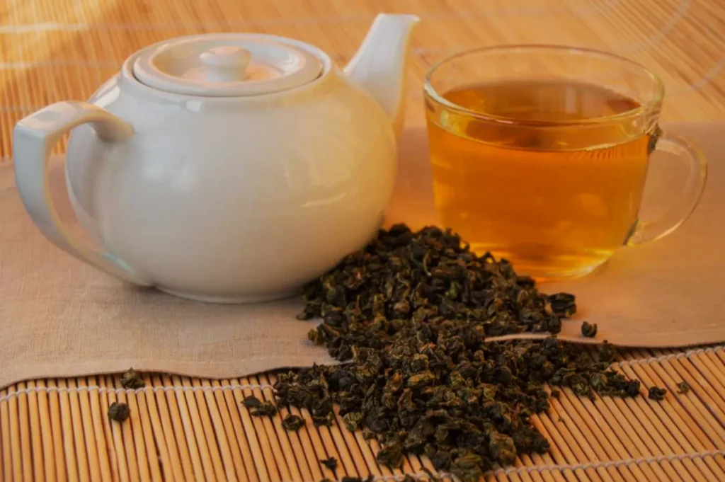 A white teapot next to a cup of tea with loose tea leaves spilled on a beige cloth.