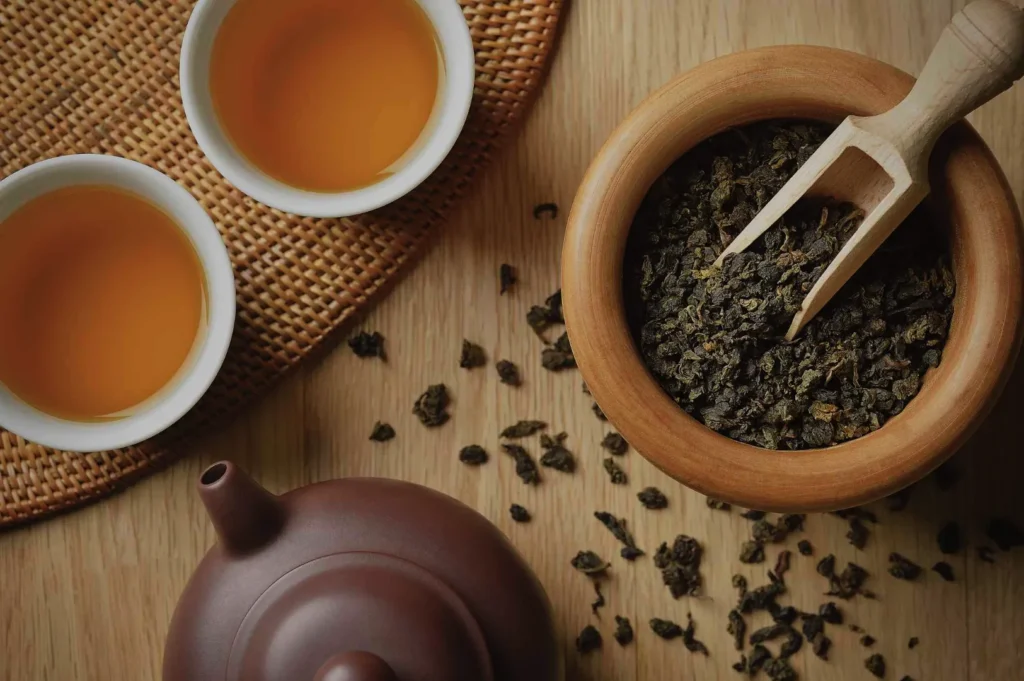 A wooden bowl filled with loose tea leaves, a wooden scooper, a teapot, and two cups of brewed tea on a wooden surface.