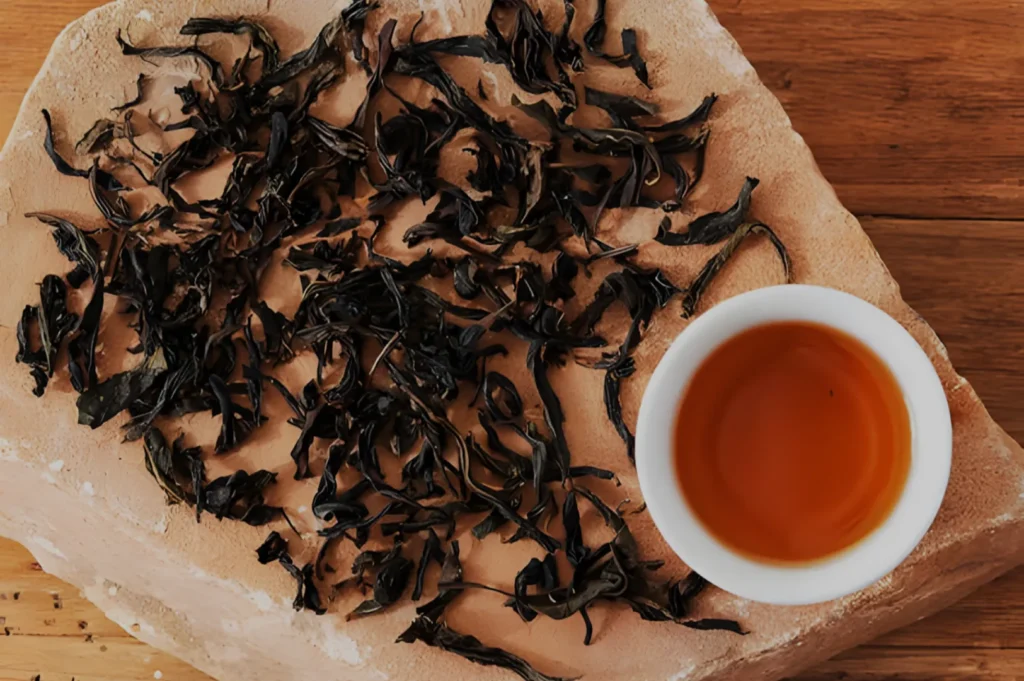 A mound of dried black tea leaves on a large rock alongside a small white cup filled with amber-colored tea.