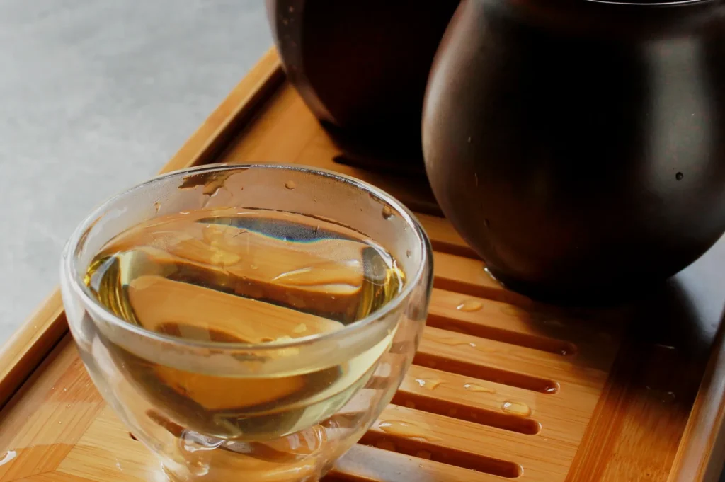 A glass cup of tea on a bamboo tray with two dark ceramic teapots in the background.