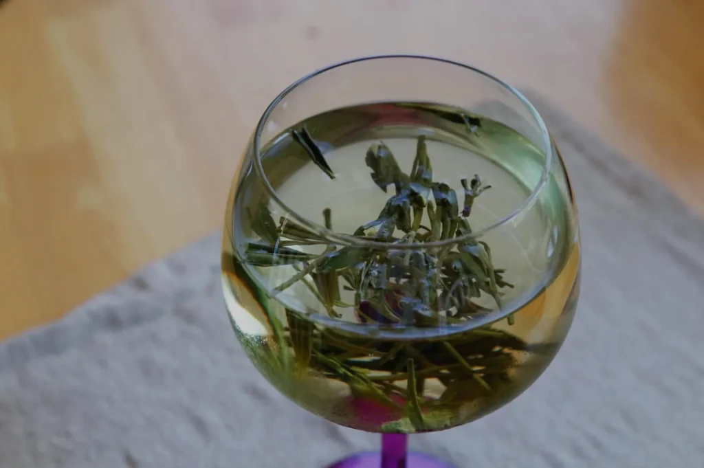 A glass of liquid with green herbs or leaves steeped inside, resting on a textured surface.