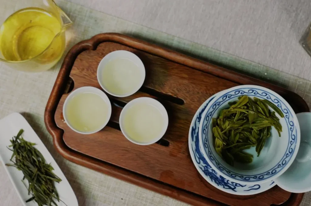 A wooden tray with three small cups filled with pale yellow tea, a bowl of green tea leaves, and a glass pitcher of tea.