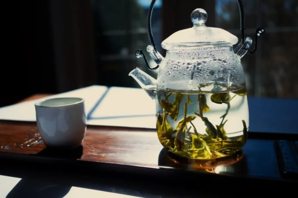 A glass teapot filled with steeping green tea leaves, placed on a wooden tray alongside a small white teacup. Bright natural light illuminates the scene.