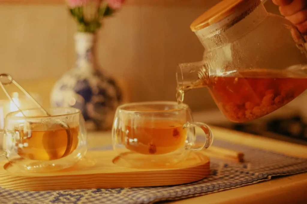 A teapot pouring tea into two glass cups on a wooden tray, with a flower vase in the background.