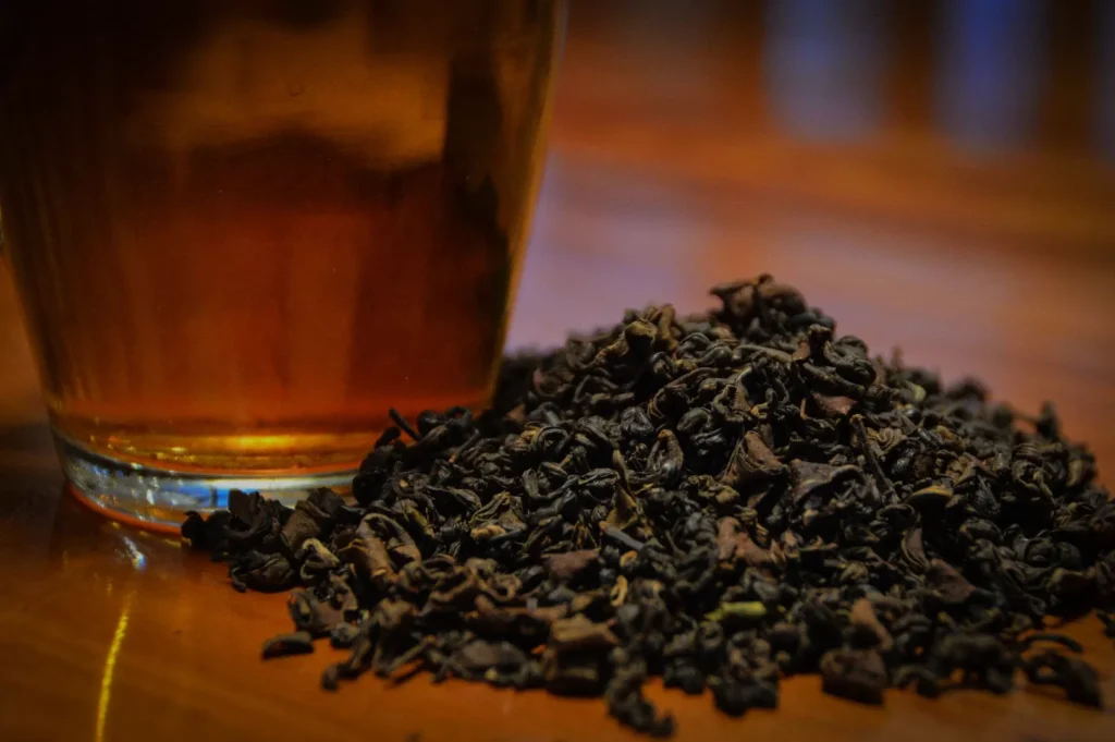 A glass of amber liquid next to a pile of loose black tea leaves on a wooden surface.