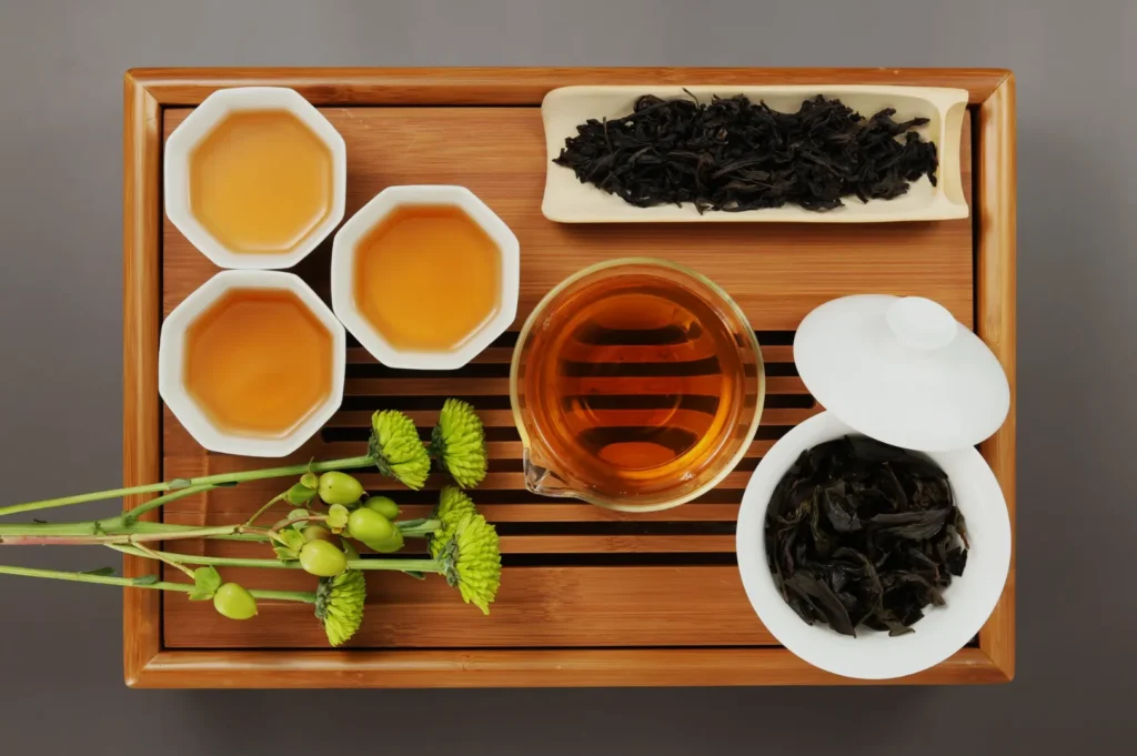 A wooden tea tray displaying four small cups of brewed tea, a glass teapot, loose tea leaves, and some green tea accessories.
