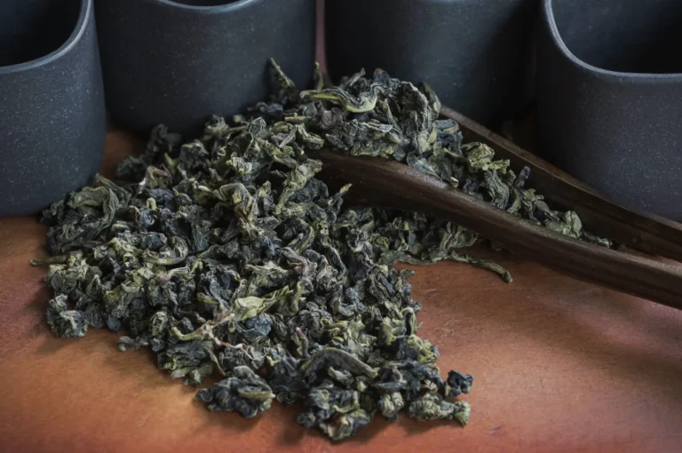 Loose dark green oolong tea leaves spill across a wooden table beside bamboo tongs and black ceramic cups, evoking a calm tea ritual.