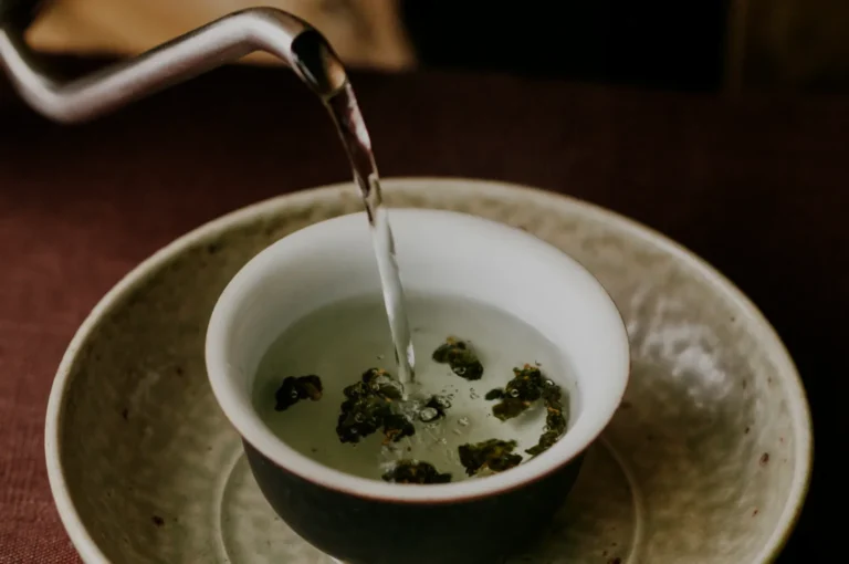 A teapot pouring hot water into a cup containing loose green tea leaves.