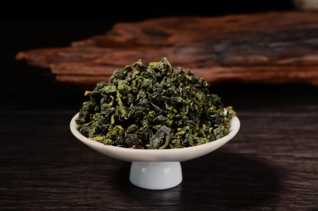 A white pedestal bowl brims with curled green tea leaves on a dark wooden table, with rustic bark behind creating a warm, earthy mood.