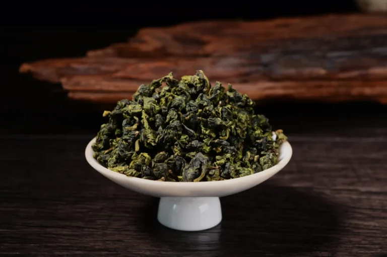 A white pedestal bowl brims with curled green tea leaves on a dark wooden table, with rustic bark behind creating a warm, earthy mood.