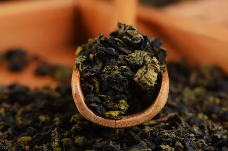 A wooden scoop filled with dried loose tea leaves, with more leaves scattered in the background.