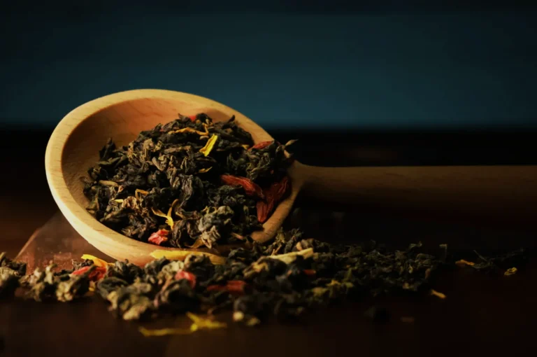 A wooden spoon filled with loose dried herbs and spices, with some scattered around it on a dark surface.