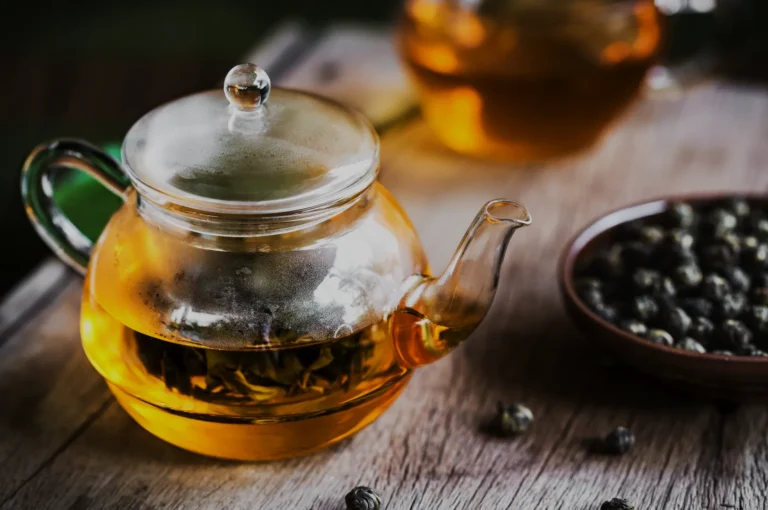A transparent teapot filled with golden tea, sitting on a wooden surface, with tea leaves visible inside. In the background, another teapot and a small bowl of loose tea leaves are present.