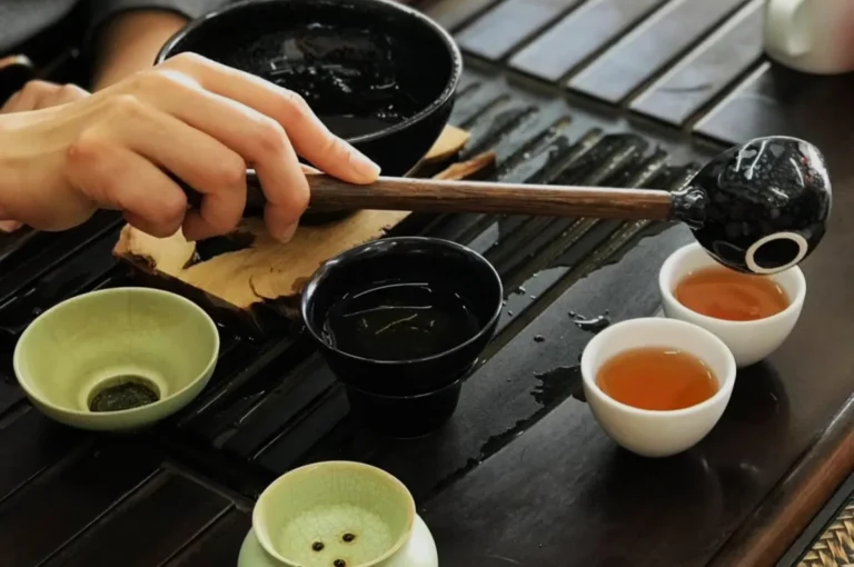 A hand holding a wooden stick hovering over a black bowl, with multiple small bowls of liquid on a wooden table.