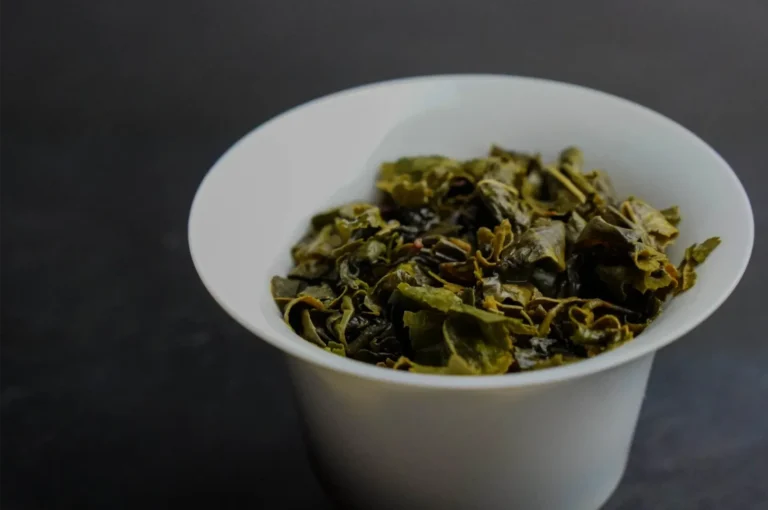 A white ceramic cup filled with loose dried green tea leaves sits on a dark surface, photographed close-up in soft light; the mood is calm and simple.
